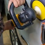 A man uses an electric buffer to polish and clean a white car door in a garage setting.