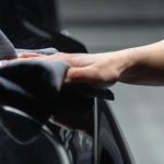 Close-up of a person's hand cleaning a shiny black car with a microfiber cloth in a garage.